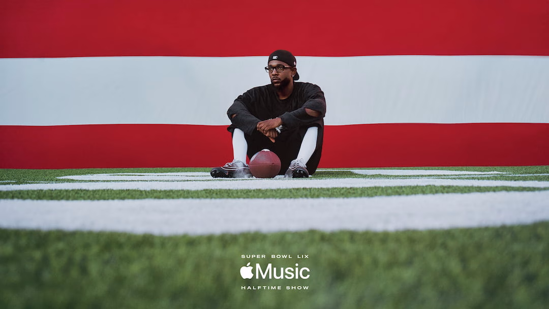Rapper Kendrick Lamar sits on a football field with an American flag behind him.