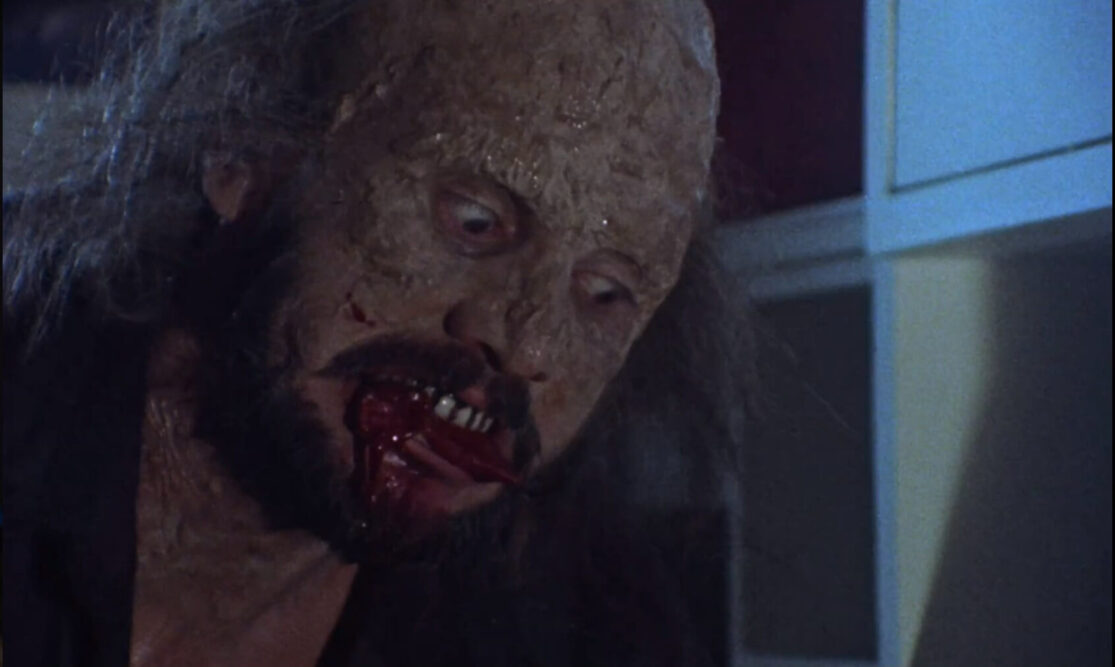 A deranged man with a heavily scarred face chews on something bloody.