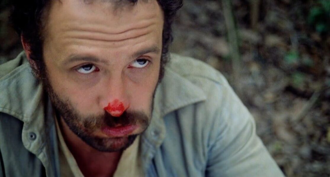 A man with a bloody nose looks up at someone unseen while chewing on something he appears not to like.