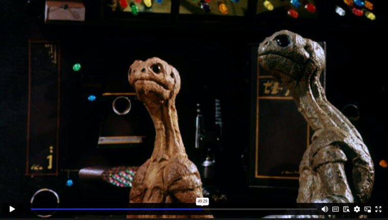 Two aliens watch a screen on the control deck of a spaceship.