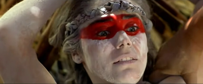 A fearful woman, her body painted powdery white except for a red band across her eyes, tied to a tree.