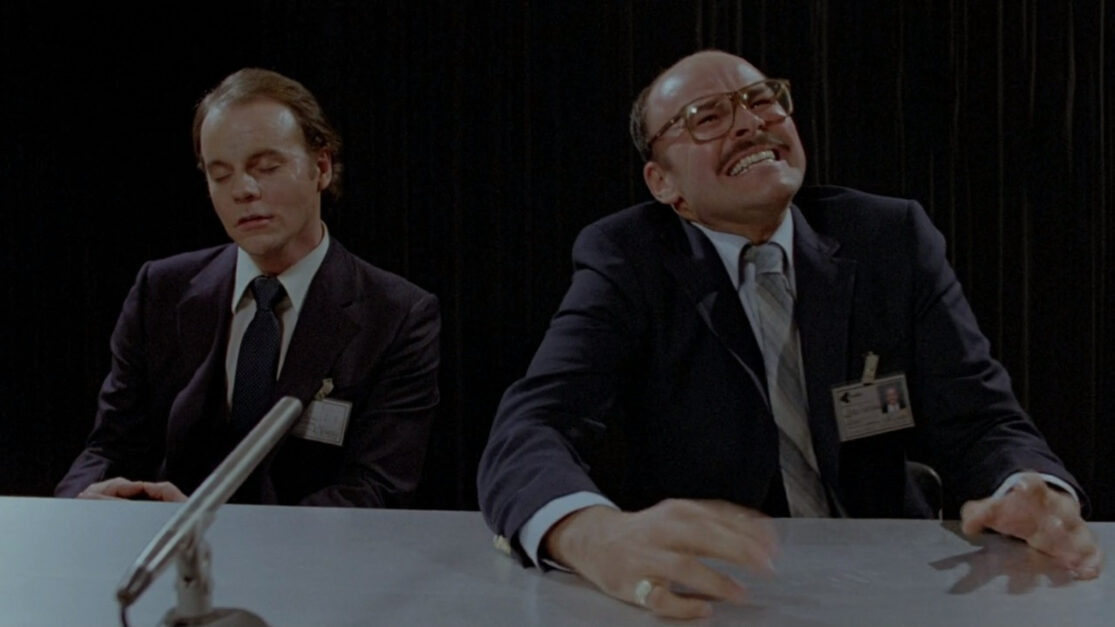 Two men sit at a conference table. One is perfectly calm, eyes closed. The other looks like his head might explode.