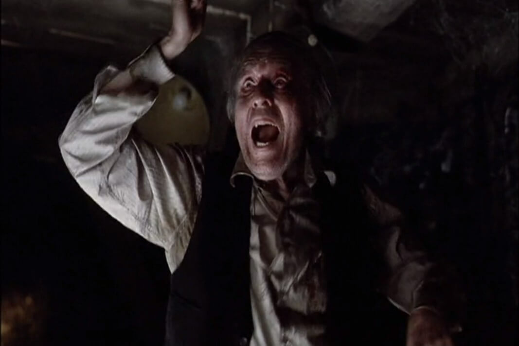 A wild-eyed, grizzled old man in dirty clothing dances and raves in a dark room.