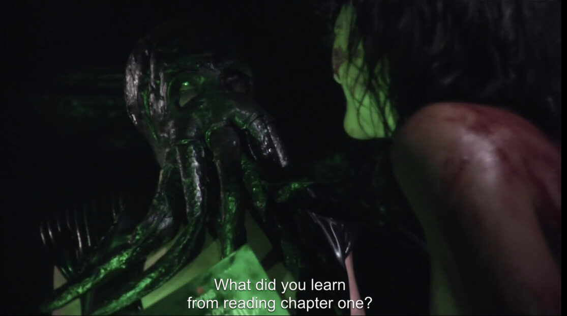 A man wearing a tentacled Cthulu mask glares at a woman, asking (in subtitles), "What did you learn from reading Chapter One?"