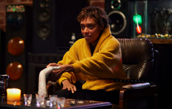 Talk to Me 2 A young woman in a yellow sweater sits in a chair. She holds a ceramic hand on the coffee table before her.