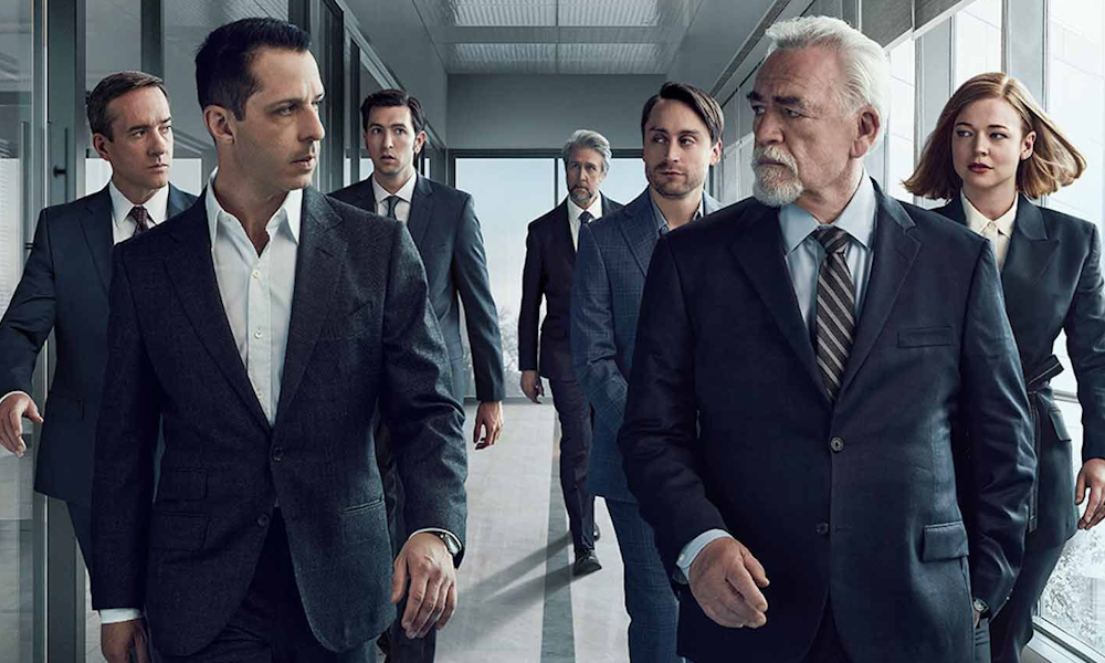 Succession1000x600 The Roy family, seven business people in suits, walking tensely in two rows down a hallway.
