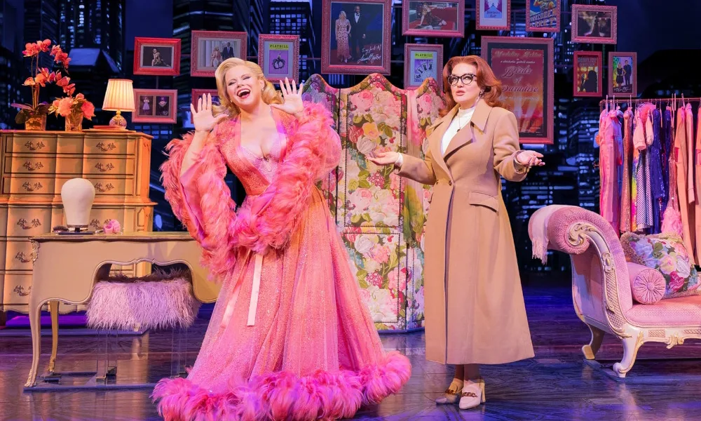 A blonde woman poses in a fancy pink robe while a red-haired woman in a trenchcoat and glasses shrugs next to her.