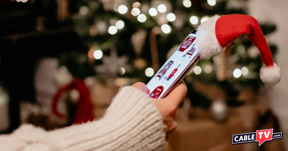 Hand holding a mobile phone that has a Santa hat on the corner. Lighted Christmas tree in the background.