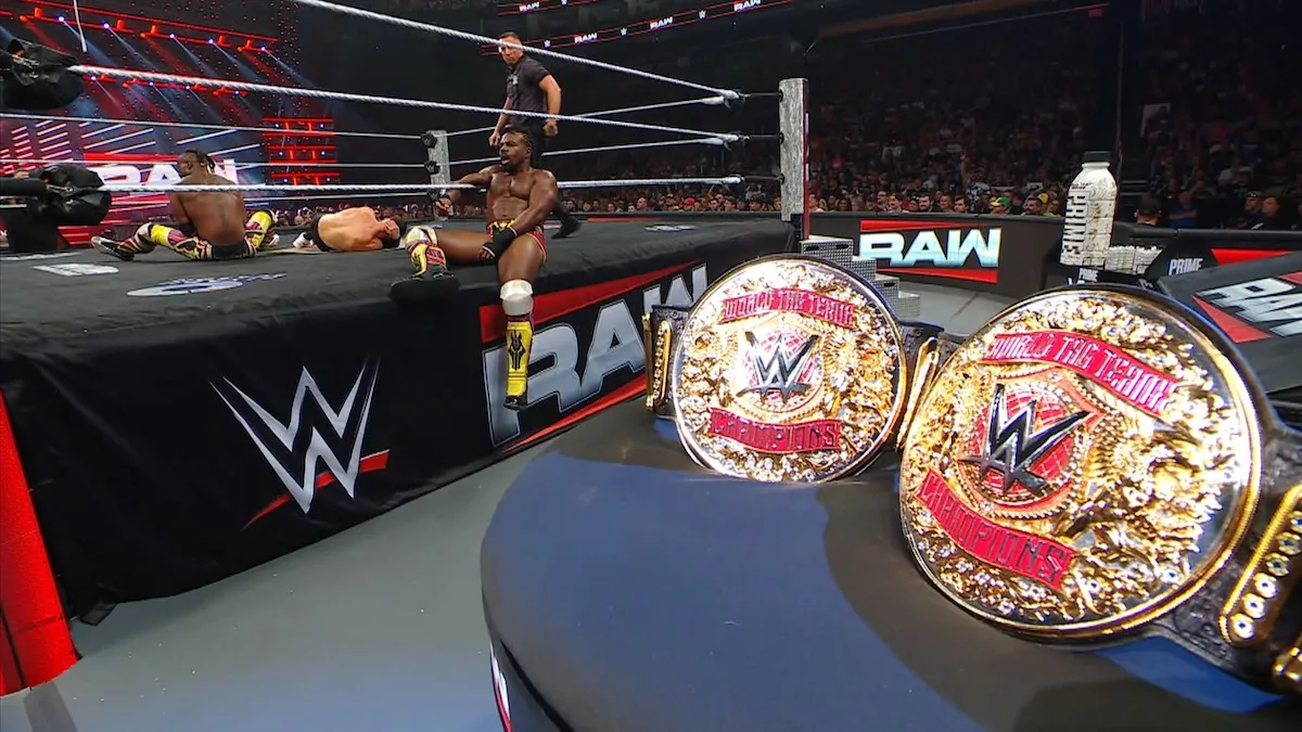 Netflix_WWE Two WWE championship belts in the foreground, with a wrestling ring and three wrestlers in the background.