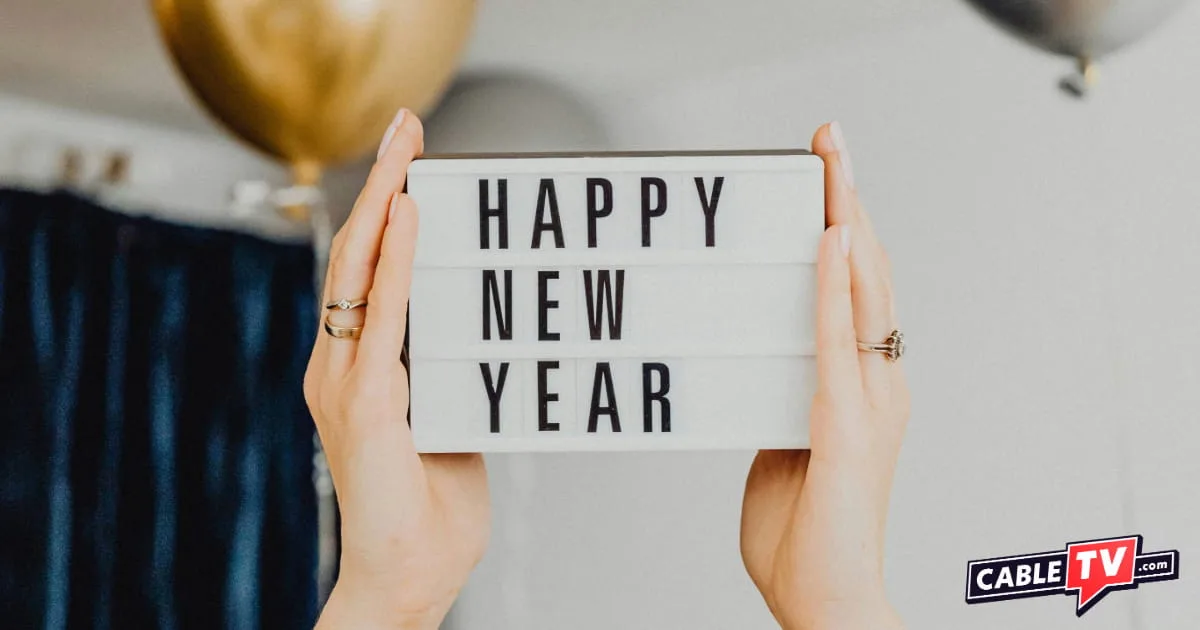 Hands hold up a small "Happy New Year" sign against a festive background of silver and gold balloons, with the CableTV.com logo in the corner.