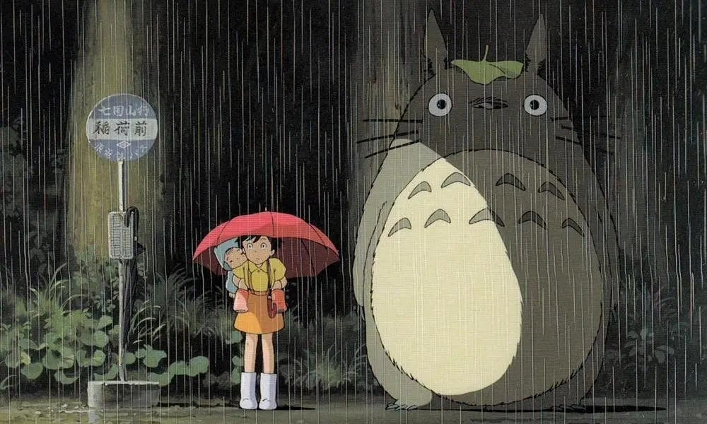 Totoro1000x600 Totoro, a large grey furry creature with a leaf on his head, stands next to a little kid in the rain.