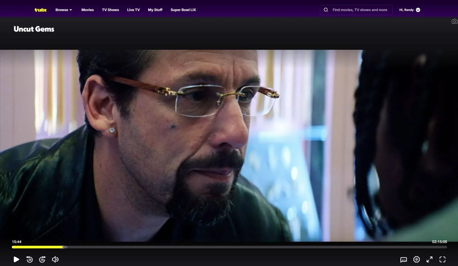 Image of a man in glasses (Adam Sandler) staring hard at a business partner in Uncut Gems, now streaming on Tubi. The image is a screenshot meant to show Tubi's user interface and controls.